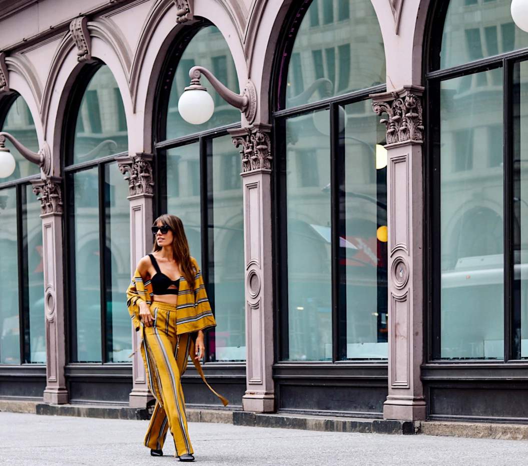 stylish influenster woman, walks strongly down a manhattan street newar 14th street in SOHO. woman wears a bright yellow two set outfit with dark shades.