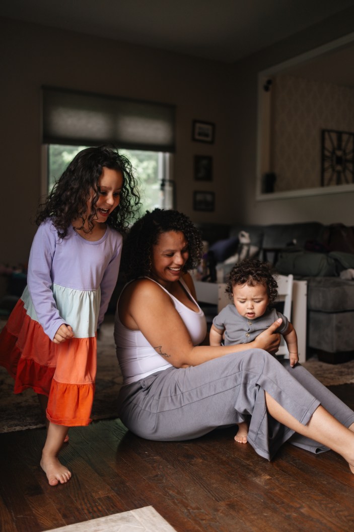 Mom holding her baby close with a warm smile while sitting on the living room floor while her toddler dances circles around her.