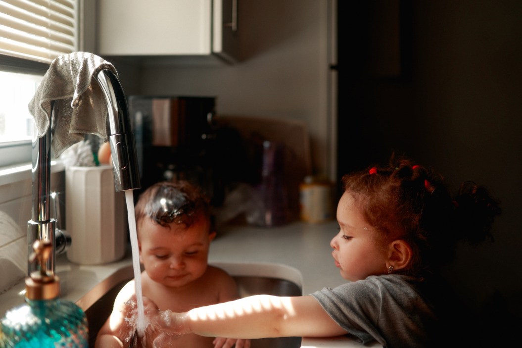 baby in kitchen sink being bathed by big sister  in moody window lighting, in stratford ct